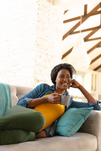 Young African American woman enjoying a moment of relaxation at home, holding a coffee cup while reclining on a sofa. Ideal for use in lifestyle blogs, home decor advertisements, or wellness articles focusing on relaxation and comfort.