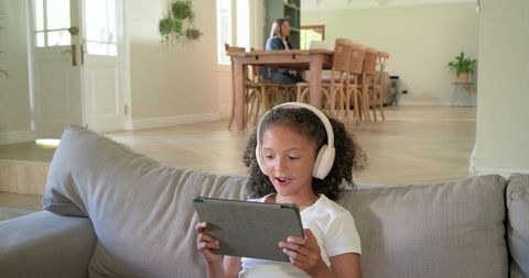 Child using tablet and wearing headphones on sofa with parent working in background
