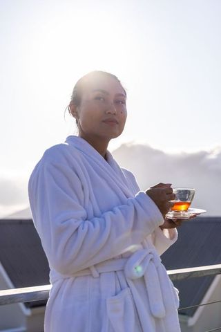 Rooftop morning calm asian woman in white bathrobe holding glass teacup during golden hour