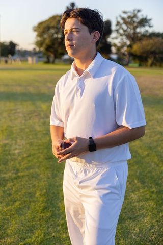 Young cricketer standing on sunlit field holding ball at golden hour wearing white kit