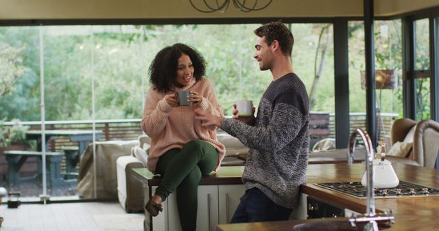 Happy diverse couple talking and drinking coffee in kitchen, woman sitting on counter. spending free time together at home.