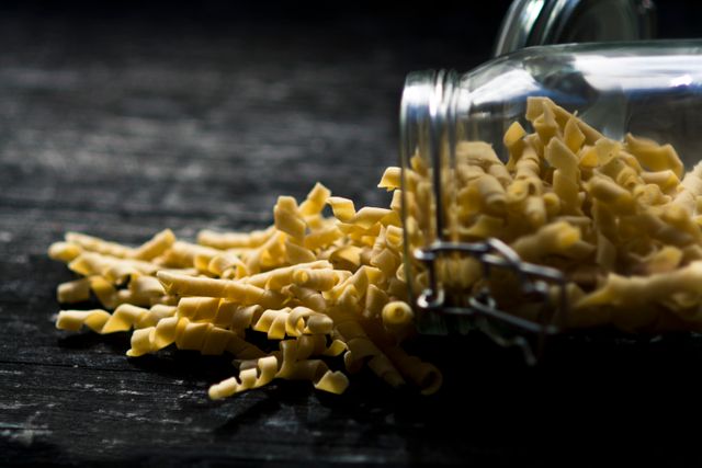 Uncooked pasta noodles spilling from a glass jar onto a dark wooden table. Detailed texture and rustic setting make this image suitable for food blogs, cooking websites, recipes, or kitchen decor. Ideal for illustrating traditional cooking ingredients or promoting authentic Italian cuisine.