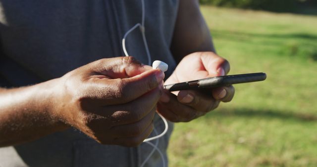 Person engaging with technology by connecting earphones to smartphone in natural environment. Useful for illustrating activities like leisure, connectivity, outdoor technology use, and wireless audio experience.