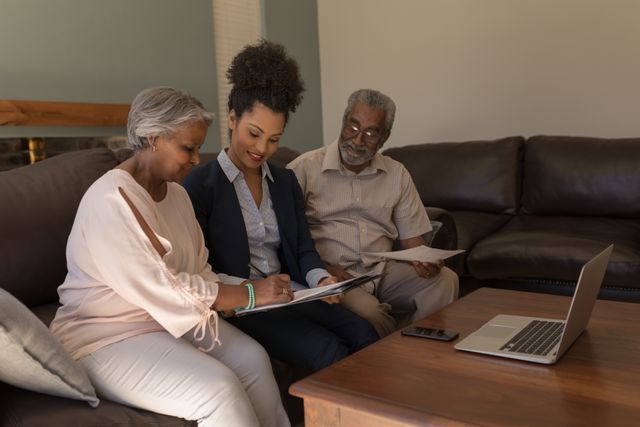 Senior African American couple sitting on sofa in living room, signing property contract with real estate agent. Ideal for use in real estate, financial services, elderly care, and family consultation contexts. Highlights professional interaction, trust, and home environment.