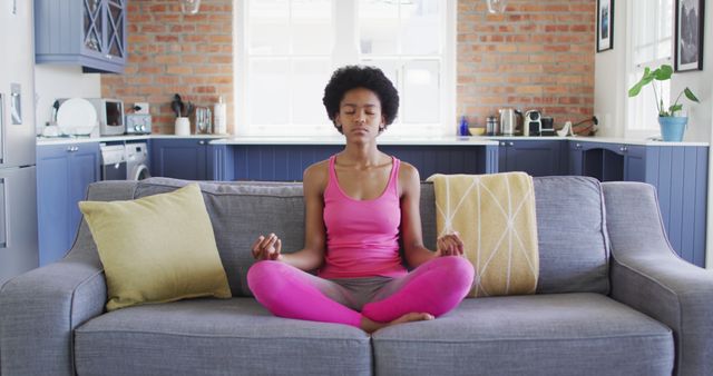 Woman with natural afro hair meditating on sofa in cozy living room setting. Wearing bright workout attire, she practices mindfulness and relaxation. Useful for content on mental health, self-care, relaxation techniques, home fitness, and lifestyle promotions.