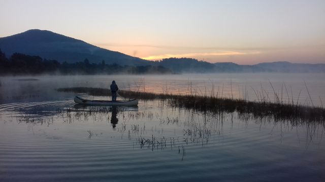 Lone fisherman standing in canoe on misty lake during sunrise, background of mountain silhouettes and still water, reflections on water, creates a tranquil and serene scene. Ideal for promoting outdoor activities, travel destinations, natural beauty, and solitude experiences.