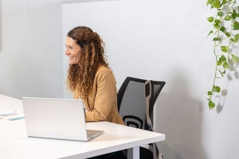 Mature professional woman smiling while working on laptop in minimalist office with greenery