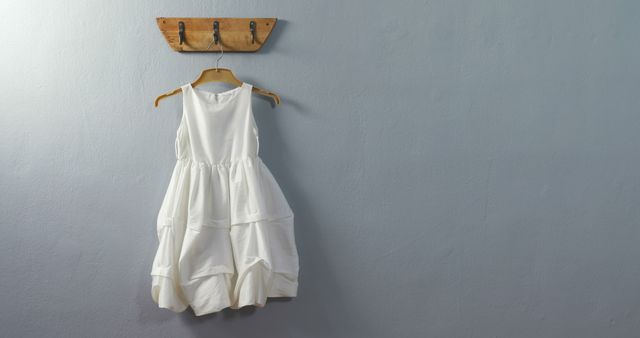 A white dress hangs on a wooden hanger against a gray wall, with copy space. The simplicity of the dress suggests a minimalist aesthetic or could indicate a special occasion like a baptism or wedding.