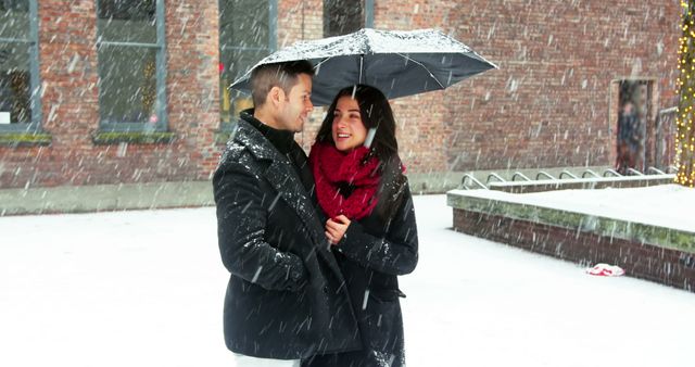 Couple standing close and sharing an umbrella during a snowy day, dressed warmly in winter clothes. Woman wearing a red scarf and black coat, both looking happy and content. Use this image for various purposes such as winter love themes, relational content, holiday marketing, romantic greetings, or seasonal promotional material.