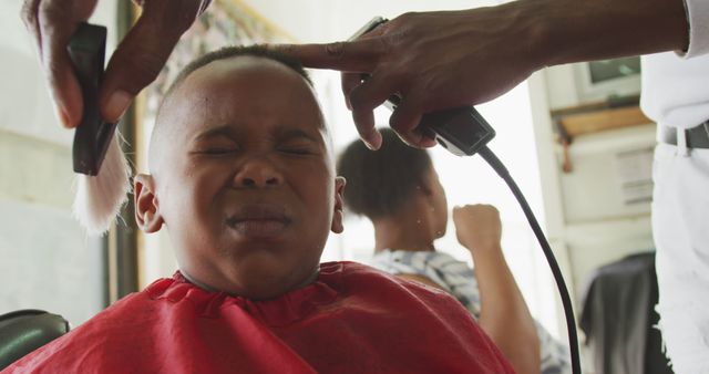 Young boy wincing while barber using hair trimmer to cut his hair. Ideal for articles on children's grooming, barbershop experiences, or lifestyle and beauty tips. Captures the reality and emotions of children undergoing routine activities.