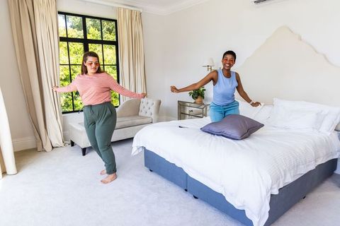 Multicultural women laughing and jumping on bed in sunlit modern bedroom
