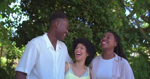 African american friends laughing under leafy canopy in sunlit park, in summer clothes