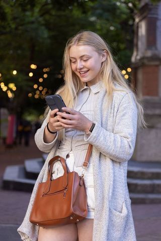 Young woman texting on smartphone in urban plaza at dusk with leather handbag