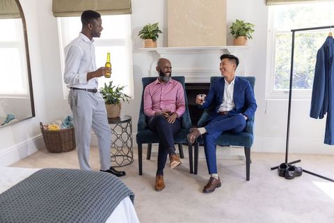Groomsmen chatting and laughing over drinks in bright dressing room before wedding ceremony