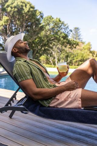 Mature african man relaxing poolside wearing white hat pink shorts holding cocktail glass