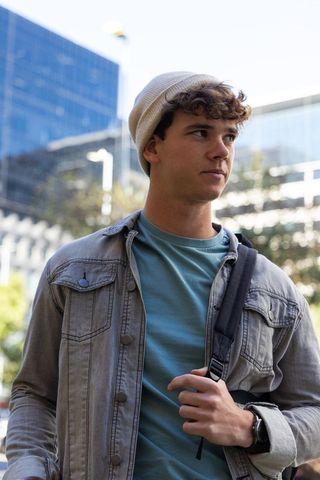 Young man wearing beanie and denim jacket carrying backpack in modern urban plaza