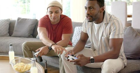 Diverse male friends sitting on sofa playing console games with controllers and snacks