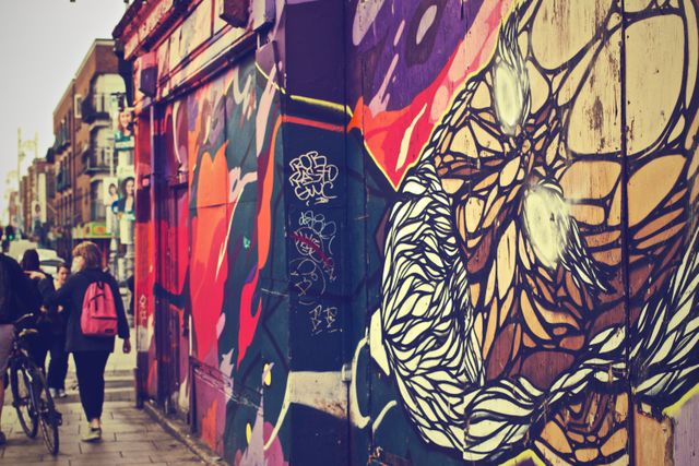 Colorful urban street art scene featuring vibrant graffiti murals on building walls, with pedestrians walking and cyclists in view. Ideal for use in digital media, websites, and marketing materials related to art, urban culture, creativity, and city life.