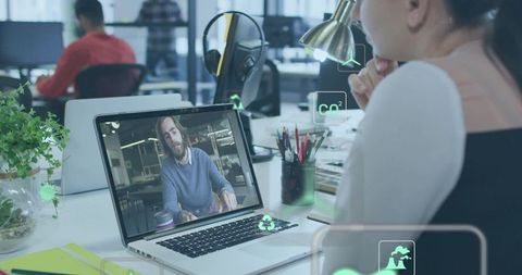 Woman watching virtual meeting on laptop at modern open-plan office with green icons