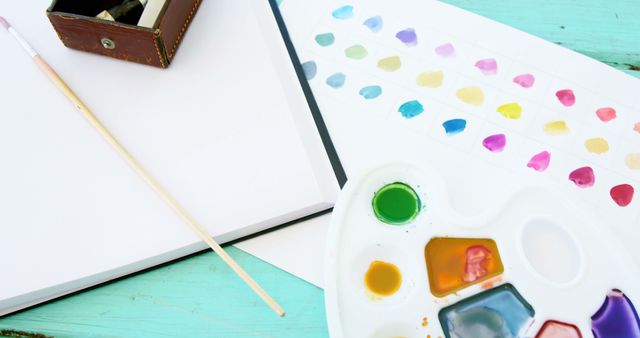 A variety of watercolors are arranged on a palette next to a blank canvas and paintbrush, with copy space. Art supplies like these invite creativity and are essential for artists and hobbyists to express their imagination.