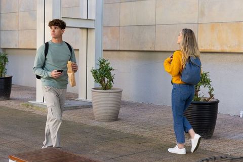 Young commuters walking through modern urban courtyard carrying backpacks, phone, paper bag