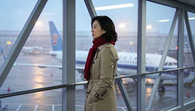 Woman waiting at airport gate in trench coat and red scarf watching plane through rainy window