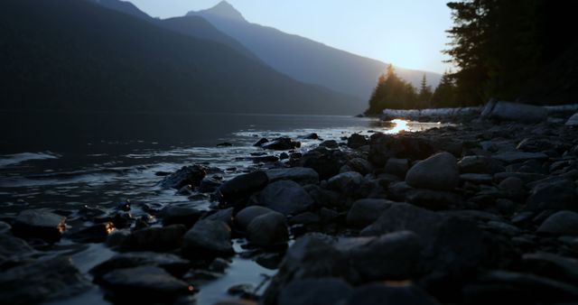 Sunlight filters through a mountainous landscape, casting a serene glow over a rocky lakeshore - Download Free Stock Photos Pikwizard.com