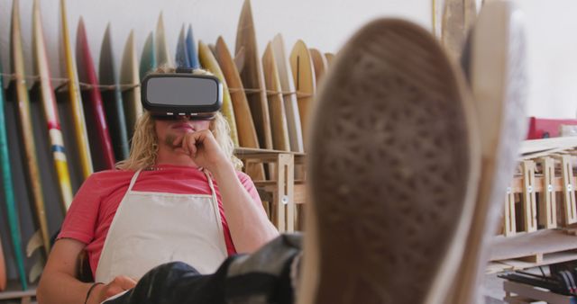 Happy caucasian male surfboard maker with feet up wearing vr headset at workshop, unaltered. Small business, technology, virtual reality, sports equipment and craftsmanship.