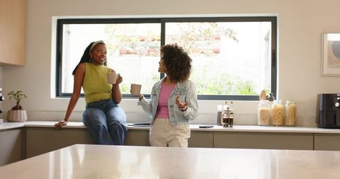 Two diverse women chatting over coffee in modern bright kitchen with large window