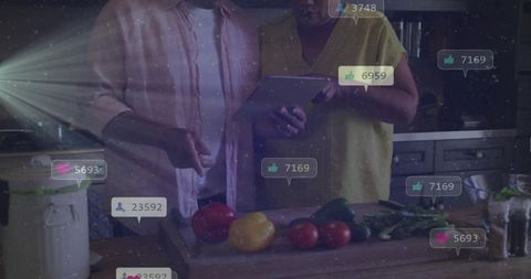 Senior couple using tablet while chopping peppers on wooden board with social media likes