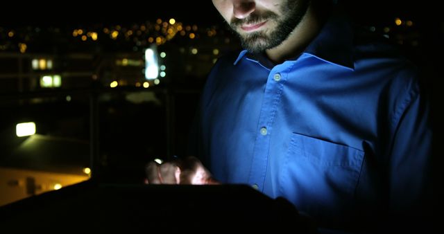 This depicts a man in an illuminated shirt using a tablet at night with city lights softly blurred in the background. Excellent for illustrating themes related to nighttime productivity, business, technology, and modern professional lifestyles. Suitable for use in articles, blogs, and presentations about working late hours, tech reliance, or digital devices.