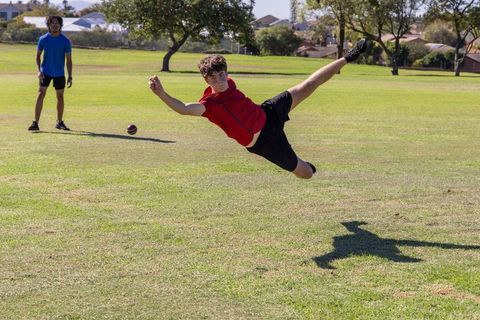 Teenager diving to catch ball on sunny park field while african american man watching