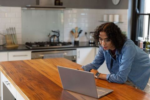 Non-binary person leaning on wooden island working remotely on laptop in modern kitchen