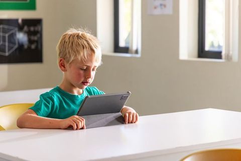 Blond boy using tablet at white desk in bright classroom learning with digital device