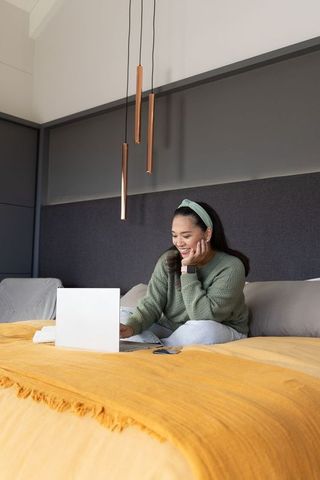 Asian woman working on laptop in cozy modern bedroom with mustard throw and pendant lights