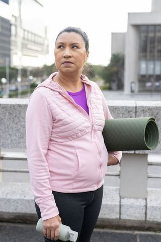 Mature woman carrying yoga mat and water bottle in urban plaza wearing pink hoodie