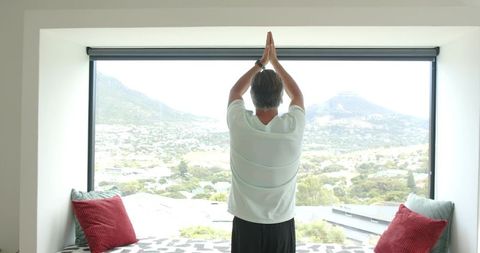Mature man stretching on window seat, practicing morning yoga with valley view, wearing watch