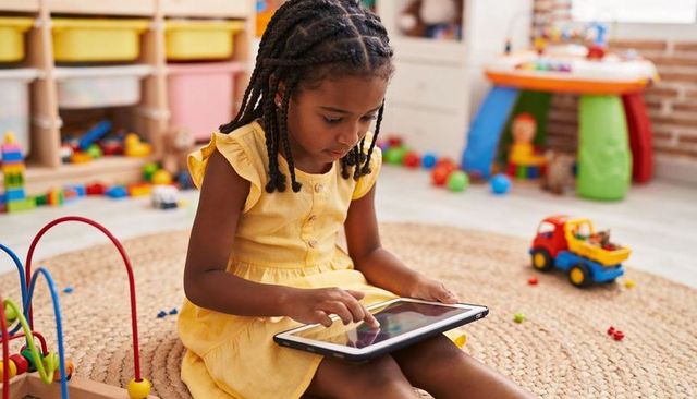 Preschool child tapping tablet on woven rug in colorful playroom for digital learning