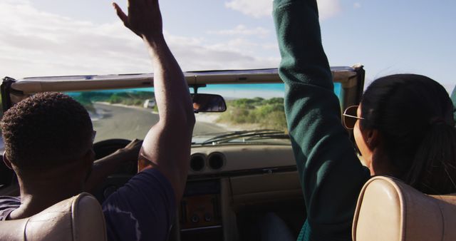Diverse couple driving on sunny day in convertible car raising hands in the air and smiling. summer road trip on a country road by the coast.