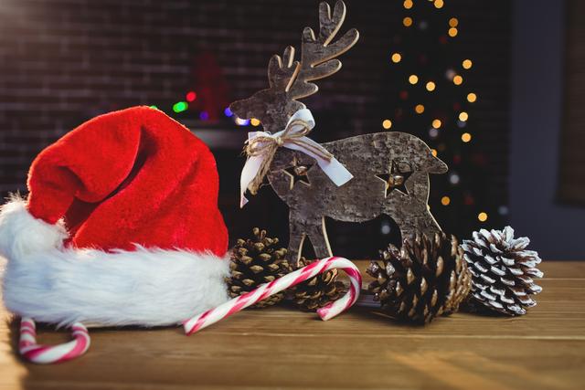 Festive Christmas decorations including a Santa hat, wooden reindeer, candy canes, and pine cones on a wooden table with blurred Christmas lights in the background. Ideal for holiday greeting cards, festive advertisements, seasonal blog posts, and social media content.