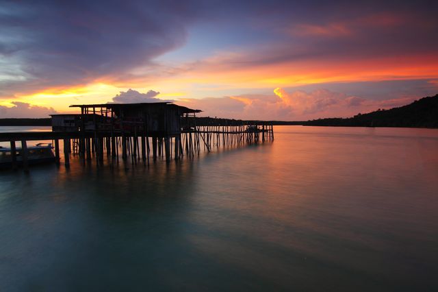 Situated over calm water, this picturesque wooden pier stretches into the horizon under a vibrant orange and purple sky at sunset. Warm, tranquil tones combined with serene reflections make this ideal for travel campaigns, relaxation and spa retreats, or backgrounds for inspirational quotes.