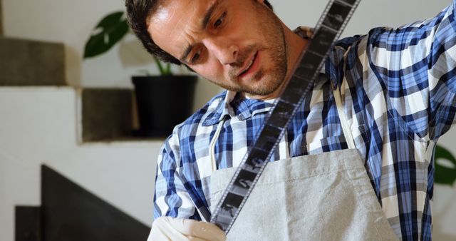 Man in plaid shirt and apron closely examining developed film strip in a home darkroom environment. Scene reflects the intricate process of traditional film photography. Ideal for use in articles or advertisements focusing on photography, creativity, or DIY projects.