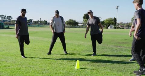 Male cricket team warming up stretching quads and sides near coach on grass field
