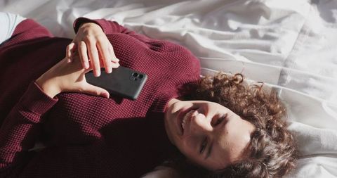 Woman Relaxing on Bed Holding Smartphone Smiling in Natural Light