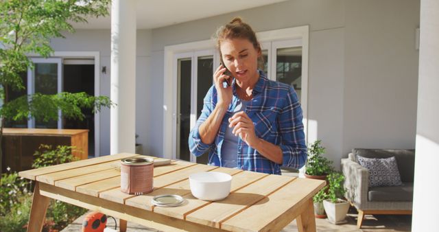 Woman talking on the phone while engaged in a DIY project. She stands near a wooden workbench outside a house, mixing paint or similar material. Could be used for topics like home improvement, multitasking, leisure activities, or residential lifestyle.