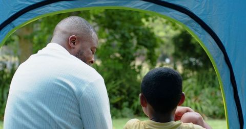 African american father and son sitting in tent on lawn bonding outdoors in green park