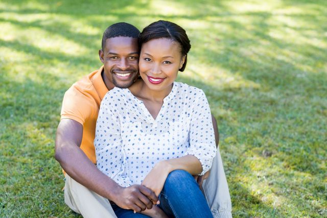 Couple sitting on grass in park, smiling and embracing. Ideal for use in advertisements, relationship blogs, lifestyle magazines, and social media posts promoting love, happiness, and outdoor activities.