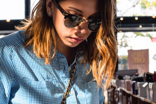 Young woman wearing sunglasses and a blue checkered shirt shopping in an indoor retail space. Lights and window in background; appears to be daytime. Suitable for use in retail, fashion, or lifestyle marketing, emphasizing modern casual style.