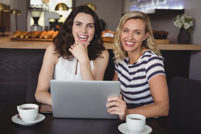 Two women sitting at a cafe table, smiling and using a laptop. They are enjoying coffee and appear to be having a good time. This image can be used for themes related to friendship, casual meetings, technology use in social settings, and leisure activities.