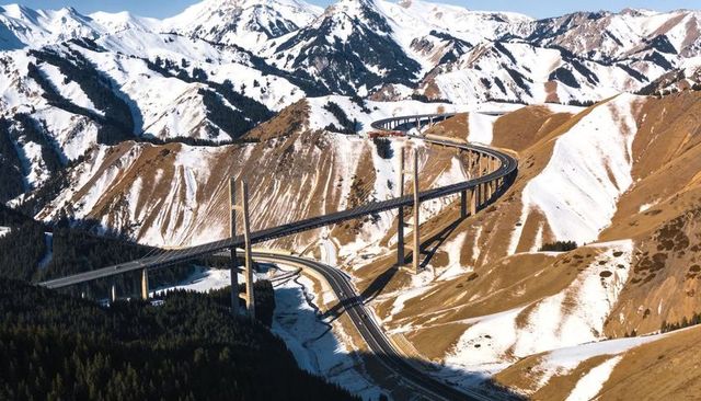 Curving viaduct crossing snowy alpine valley with tall piers and conifer forest views